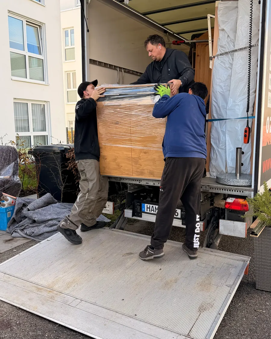 Drei Mitarbeiter heben einen in Folie verpackten Schrank auf die Ladebordwand des Umzugswagens